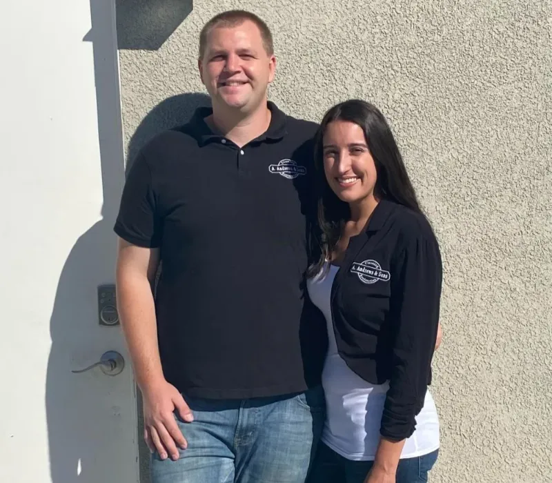 A man and woman smiling and standing side by side against a textured wall, both wearing black tops.