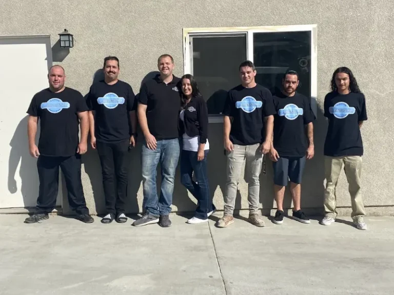 Seven people stand in a row outside against a beige wall, most wearing black shirts with a blue logo.