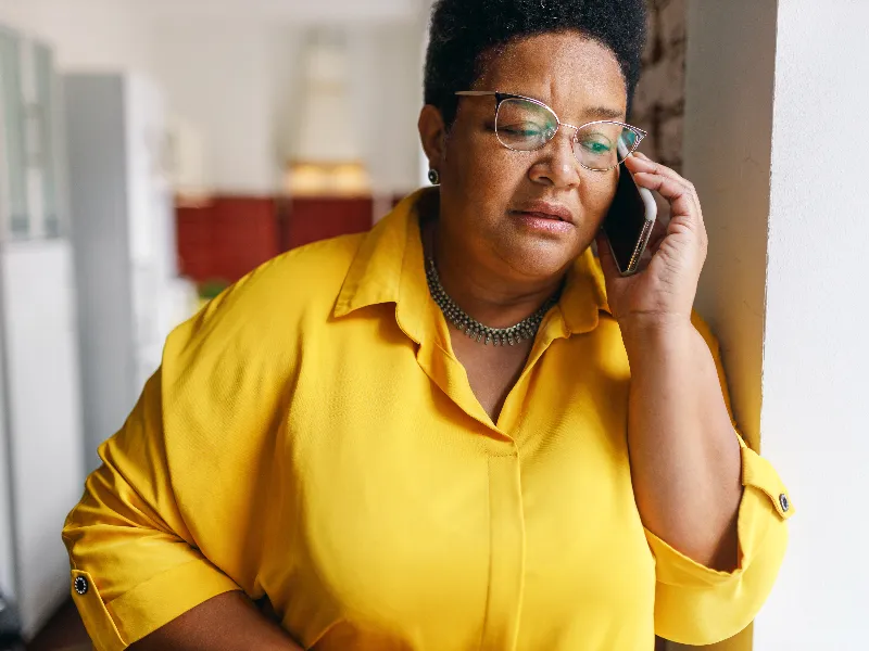 Woman in yellow shirt leaning against a wall, talking on a phone with a concerned expression.