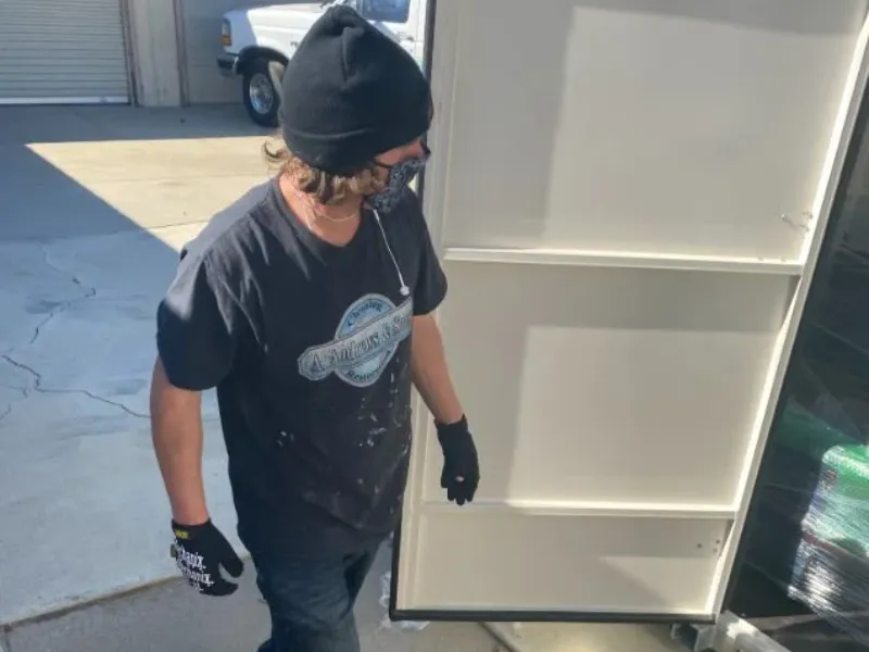 Man wearing a black beanie, gloves, and mask standing next to a large white panel outdoors.