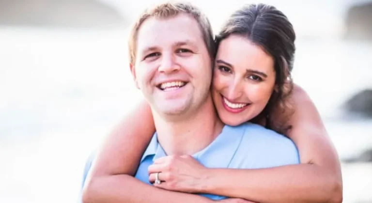Smiling woman hugs smiling man from behind, both looking at the camera.