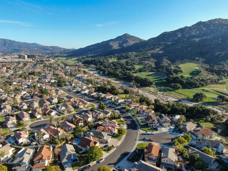 Suburban neighborhood with houses and winding roads next to green hills under a clear blue sky.