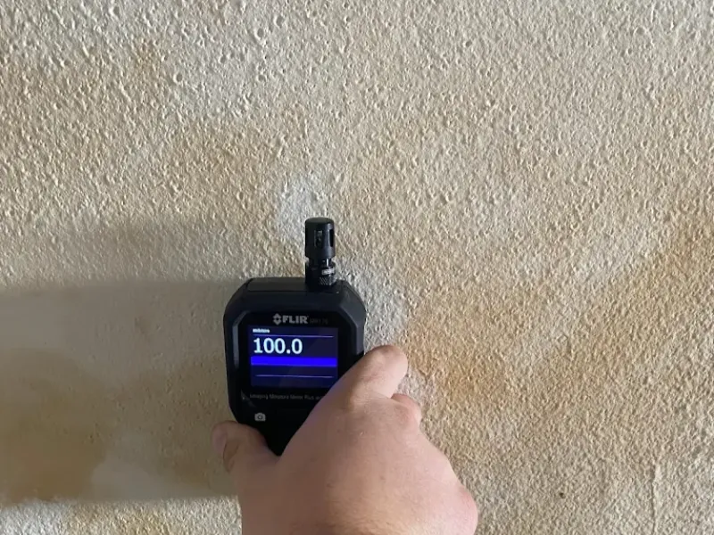 Hand holding a FLIR gas detector showing a reading of 100.0 against a textured wall.