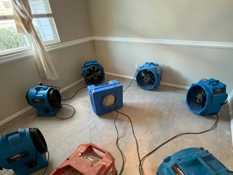 Blue air movers and a red dehumidifier arranged in an empty room with bare floors.