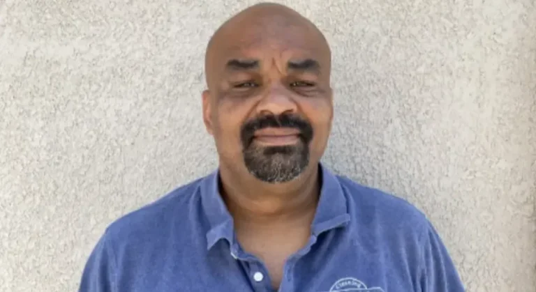 Man with a goatee wearing a blue collared shirt stands against a textured light wall.